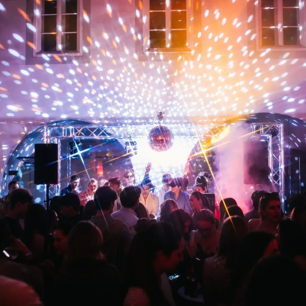Menschen tanzen auf Party mit Lichteffekten und Nebel unter Spiegelkugel.
